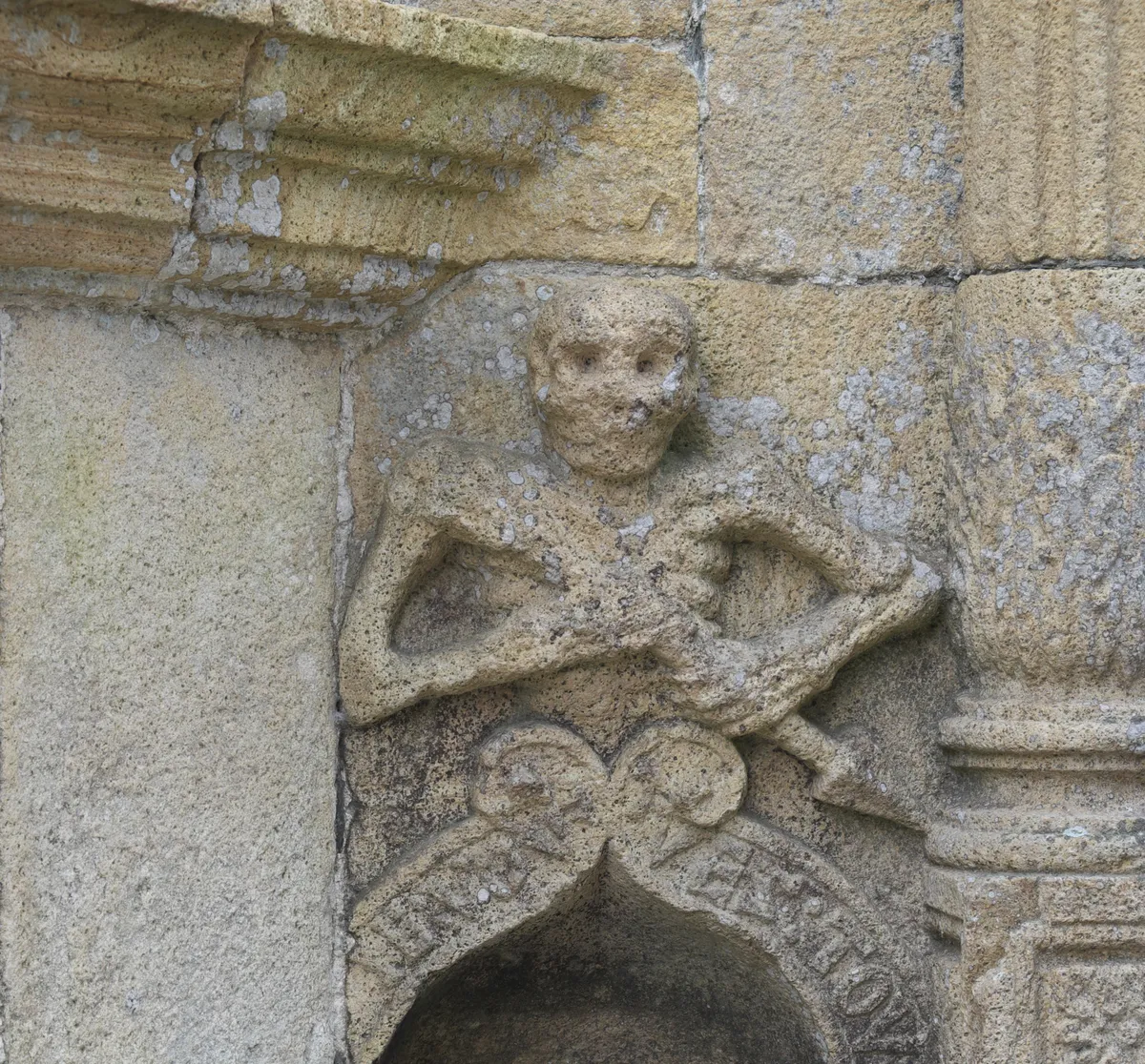 L'Ankou sculpté au-dessus du bénitier de l'ossuaire de La Roche-Maurice, un squelette qui tient une flèche, au-dessus de l'inscription « Je vous tue tous »