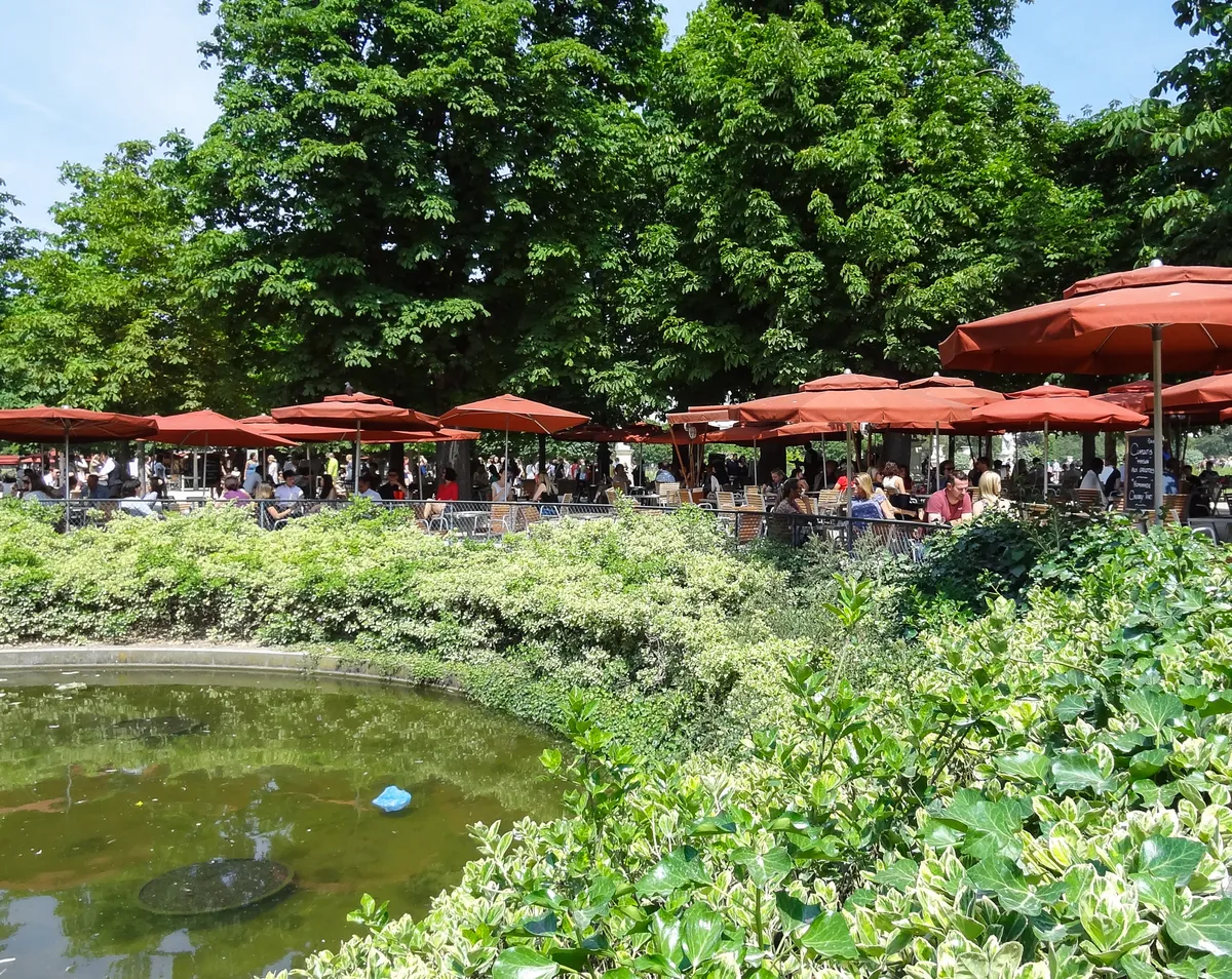Les parasols rouges d'une terrasse sous les arbres du jardin des Tuileries, derrière un bassin et des massifs