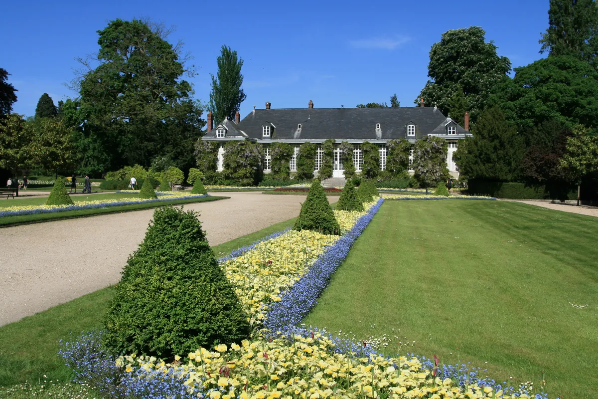 Des parterres à la française, buis taillés et massifs fleuris, devant un pavillon au toit d'ardoise
